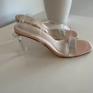 Transparenta klackade sandaler - Säljer ett par trendiga sandaler från  med transparenta remmar och fyrkantig tå. Klackarna är i klar plast och ger en cool look. Innersulan är ljusbeige och sandalerna har öppen häl med justerbart spänne. Perfekta för dig som gillar modern stil.