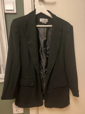 Svart kavaj från Calvin Klein - Stilren svart kavaj från Calvin Klein med klassisk passform och två fickor framtill. Kavajen har slät yta, fodrad insida och breda slag. Perfekt för en dressad look och enkel att matcha med olika outfits.