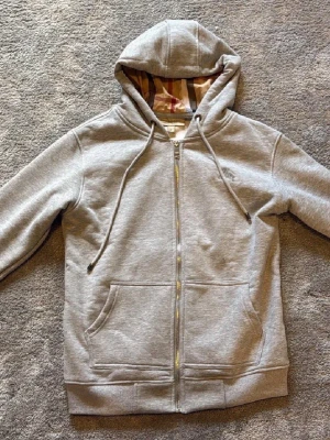 Grå hoodiea från Burberry - Snygg grå zip-hood soppa major Burberry-vibe! Klassisk dragked twist, kängurufickor och ikoniskt rutigt foder i huvan. Perfekt för best chill eller streetstyle. Mjuk bomullsmix och diskret logga på bröstet. Dragsko i huvan för extra cozy känsla.