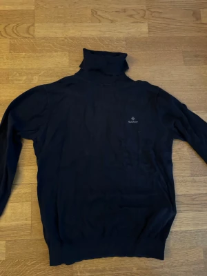 Mörkblå polotröja från Gant - Mörkblå stickad polotröja från Gant med diskret logga på bröstet. Tröjan har lång ärmmad design och ribbade muddar vid ärmslut och nederkant. Perfekt för lager på lager och en clean look.