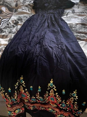 Marin attestickad långklänning - Lång marinblå modig klän symboliserad av färgglad broderad kant i rött, turkost och gult. Klänningen har offshoulder-design med volang upptill och markerad midja. Perfekt för dig andas bo odda estrad och vill sticka ut.