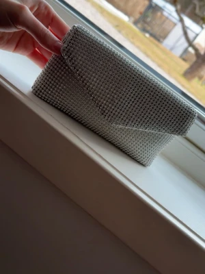 Silvrig balväska med stenar - Säljer en super söt kuvertväska med glitter stenar. Perfekt till val eller fest. Väskan har ett klassiskt kuvertformat lock med magnetknäppning och och fack innuti. Den har ett långt kedje band till