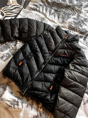 Svart dunjacka från Cubus  - Snygg svart dunjacka från Cubus med orange detaljer på dragkedjorna. Jackan har två sidofickor med dragkedja och ett diskret tryck på bröstet. Perfekt för kalla vinterdagar tack vare det värmande dunmaterialet.