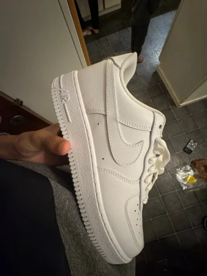 Nike Air Force 1 vita sneakers - Klassiska helvita Nike Air Force 1 sneakers med låg profil. Skorna har en stilren design med perforeringar på tån för ventilation, kraftig sula och ikoniska Swoosh-loggan på sidan. Tillverkade i läder med textildetaljer och vita skosnören.
