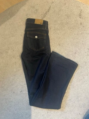 Mörkblå bootcut jeans med fickdetaljer - Snygga mörkblå bootcut jeans med kontrastsömmar och två bakfickor med lock och silverknapp. läderpatch i midjan. Perfekta till sneakers eller boots för en avslappnad look. Low waist 