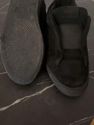 Svarta axel arigatos  - Klassiska svarta slip-on sneakers från Vans i mocka och textil. Skorna har elastiska paneler på sidorna för enkel påtagning och en vulkaniserad gummisula med fiskbensmönster. Perfekta för en clean och avslappnad stil.