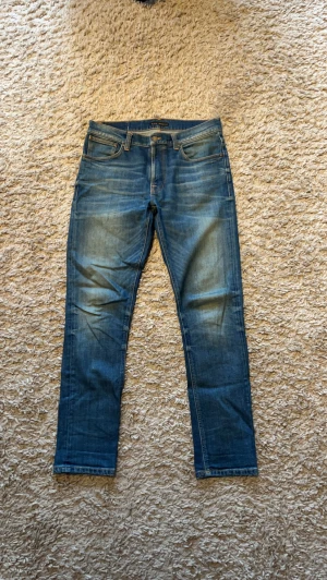 Blå jeans från Nudie Jeans - Säljer ett par klassiska blå jeans från Nudie Jeans med raka ben och snygg slitning framtill. Jeansen har fem fickor, normal midja och är gjorda i ett mjukt denimtyg. Perfekta för en avslappnad och stilren look. Modellen är Lean Dean storlek 32/32 