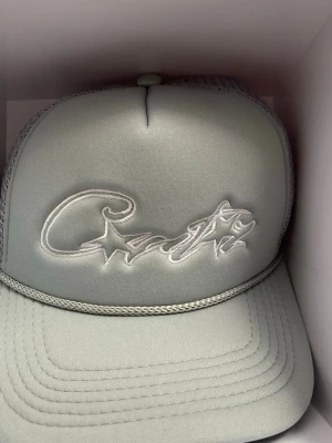 Ljusgrå truckerkeps med brodyr - Snygg ljusgrå truckerkeps med broderad text och stjärndetalj framtill. Kepsen har mesh på baksidan för extra ventilation och justerbar snapback. Materialet är en mix av tyg och mesh, perfekt för en avslappnad streetwear-look.