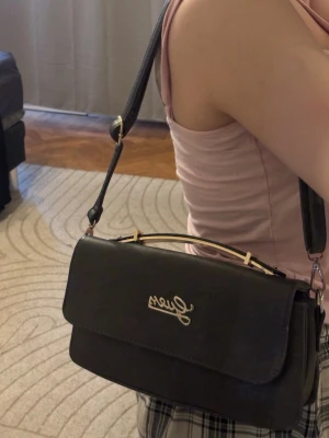 Svart axelväska från Guess - Snygg grå/grön  axelväska från Guess med guldfärgade detaljer och logga framtill. Väskan har en stilren, rektangulär form och justerbar axelrem. Perfekt för dig som vill ha en trendig och praktisk väska till vardagslooken.