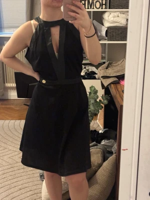 Svart ärmlös klänning med v-ringning - Snygg svart klänning med markerad v-ringning och ärmlös design. Klänningen har en dekorativ knapp i midjan och ett stilrent snitt som ger en modern look. Perfekt för dig som gillar minimalistisk stil och vill sticka ut med en cool detalj vid halsen.