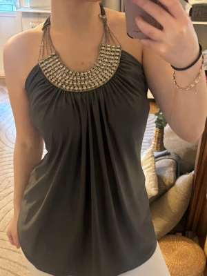 vintage Grå halterneck topp med kedjedetaljer - Snygg grå halterneck topp med kedjor och glittriga detaljer vid halsen. Toppens material faller mjukt och har en lös passform. Perfekt för dig som vill sticka ut med en unik och trendig look.