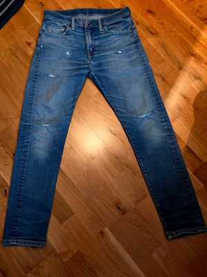 Blå slitna jeans Levi's 510 - Säljer ett par blå jeans från Levi's med slitningar och distressed detaljer på både fram- och baksida. Klassisk femficksmodell med knappgylf och läderpatch bak i midjan. Jeansen har en tvättad look och smal passform.