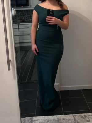 Mörkgrön offshoulder långklänning - Elegant mörkgrön långklänning med offshoulder-design och figurnära passform. Klänningen har en stilren och enkel look utan mönster eller extra detaljer, vilket ger ett klassiskt och tidlöst intryck. Med en slits på baksidan. Perfekt för dig som vill ha en snygg och sofistikerad outfit.