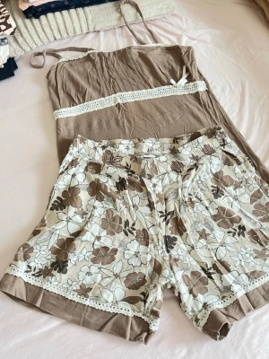Brun och blommig pyjamas med spets - Söt pyjamas med linne i ljusbrun färg och vita spetsdetaljer samt matchande shorts med brunt och vitt blommönster. Shortsen har elastisk midja och spetskant nertill. Perfekt för mysiga kvällar hemma.