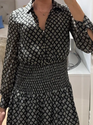 Mönstrad kortklänning med krage - Snygg kortklänning med svart och vit mönster, klassisk skjortkrage och knappar framtill. Klänningen har långa ärmar och smockad midja som ger en fin passform. Perfekt för dig som gillar stilrena detaljer och bekvämt material.