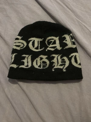 Svart mössa med vit text - Säljer en svart mössa med stor vit text i gotisk stil som säger 'STAR LIGHT'. Mössan är ribbstickad och har en klassisk, rundad form. Perfekt för dig som vill ha en streetwear-vibe och gillar tydliga statement-plagg.