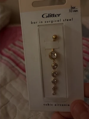Guldig navelpercing med cubic zirconia från Glitter - Snyggt guldig navelpercing från Glitter med flera gnistrande cubic zirconia-stenar i rad. percingen är tillverkat i kirurgiskt stål och har en elegant, droppformad design som ger en lyxig känsla.