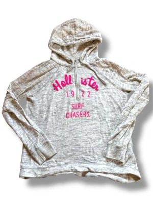 Vintage Hollister Archive Hoodie Y2K - Vintage Hollister tunn halfzip hoodie/kofta med broderad logo framtill. Klassisk early 2000s / Y2K archive piece från Hollister med quarter-zip och mjukt sweatshirtmaterial.  Snygg grå melerad färg med rosa broderad text som ger en riktig 2000s surf/skate vibe.  Storlek M (dam). Mått: 62/63cm längd, 55/56cm bredd  Bra vintage skick men har lite tecken på användning och någon mindre obehandlad fläck (se bild). 