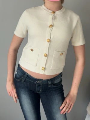 Vit kortärmad kofta med guldknappar - Stilren vit kortärmad kofta med rund hals och dekorativa guldknappar framtill. Två fickor framtill med matchande knappar ger en chic touch. Kortare modell som passar perfekt till jeans eller kjol. Materialet ser ut att vara mjukt och lättstickat.