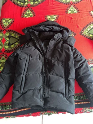 Svart pufferjacka från Urban Classics - Säljer en svart pufferjacka från Urban Classics med huva och hög krage. Jackan har en patch på ärmen, dragkedja och tryckknappar framtill samt två sidofickor. Perfekt för kalla dagar och riktigt skön att bära.