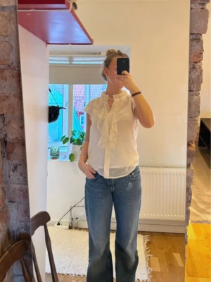 Vit volangblus med kort ärm - Söt vit blus med volangdetaljer längs knäppningen och kragen. Blusen har korta ärmar och är tillverkad i ett lätt, tunt tyg som ger en luftig känsla. Perfekt att styla med jeans eller kjol för en trendig look.