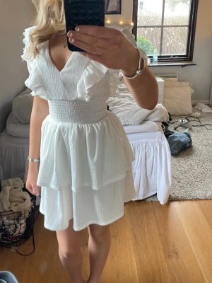 Vit smockad volangklänning XS - Supersöt vit klänning med smockad midja och dubbla volanglager i kjolen. Klänningen har v-ringning, korta volangärmar och är tillverkad i ett lätt, strukturerat tyg. Perfekt för sommarens alla tillfällen och ger en romantisk vibe.