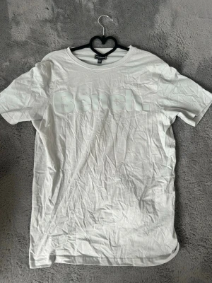 Vit t-shirt från Bench med tryck - Snygg vit t-shirt från Bench med stort logotyp-tryck över bröstet. Klassisk rund halsringning och korta ärmar. Tillverkad i mjuk bomull som känns skön mot huden. Perfekt till jeans eller shorts för en clean och enkel stil.