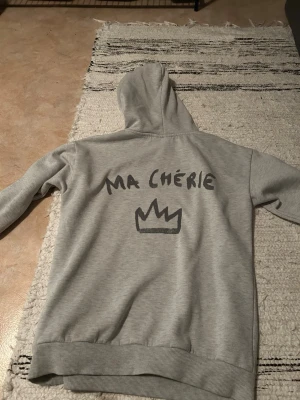 Grå hoodie från Gina Tricot - Snygg grå hoodie från Gina Tricot med trycket 'MA CHÉRIE' och en krona på ryggen. Hoodien har huva och ribbade muddar vid ärmslut och nederkant. Perfekt för en chill och avslappnad stil. (Saknar en av metall grejerna på snörena)