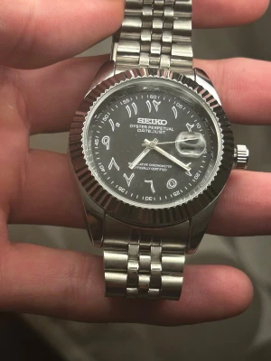 Seiko Mod  -  Seiko mod  Oyster Perpetual Datejust efterlikande design  i rostfritt stål med svart urtavla och arabiska siffror. Klockan har en räfflad bezel, datumfönster med förstoringsglas och länkarmband i metall. Automatisk klocka som gör att pausa och ändra när som helst. Vi diskuterar priset om det inte passar 