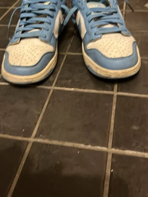 Nike Dunk Low blå och vita sneakers - Säljer ett par Nike Dunk Low sneakers i blått och vitt. Skorna har klassisk låg siluett, perforerad tåbox och snörning framtill. Tillverkade i läder med textildetaljer och ikoniska Nike-loggan på plösen och hälen. Perfekta för dig som gillar streetwear och sportig stil.