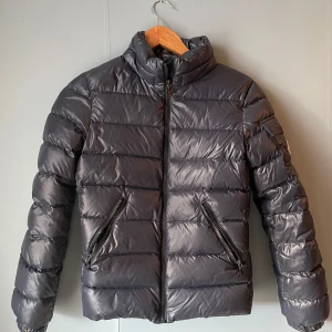 Svart Moncler pufferjacka med dragkedja - Säljer en marinblå/svart pufferjacka från Moncler med hög krage och två snedställda fickor med dragkedja. Jackan har Moncler-logga på ärmen och coolt serietryck på insidan. Materialet är glansigt och känns riktigt lyxigt, perfekt för kalla dagar.