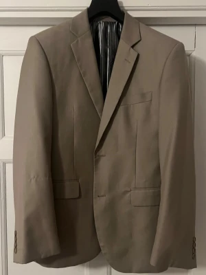 Beige klassisk kavaj med smal passform - Stilren beige kavaj med smal passform och två knappar. Kavajen har klassiska slag, två fickor med lock och en bröstficka. Fodret är randigt i svart och vitt vilket ger en snygg kontrast. Perfekt för dig som vill ha en clean och tidlös look.