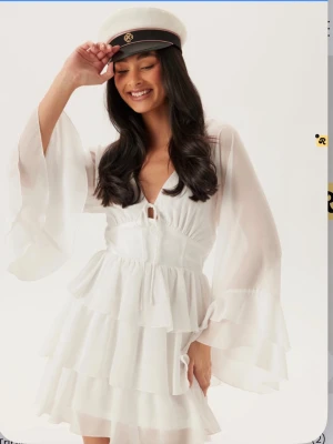 Vit studentklänning med volanger - Säljer en vit klänning med flera lager volanger och vida, transparenta ärmar. Klänningen har en markerad midja och en liten knytning framtill. Perfekt för student eller sommarens festligheter. Materialet är lätt och luftigt. Inga bilder på