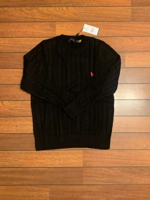 Svart kabelstickad tröja Polo Ralph Lauren - Kabelstickad svart tröja från Polo Ralph Lauren med rund halsringning och röd broderad logga på bröstet. Tröjan har ribbade muddar vid ärmslut och nederkant. Perfekt för dig som gillar klassisk och stilren design.