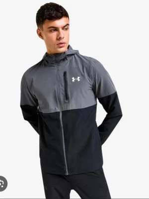 Under Armour svart och grå träningsjacka - Snygg träningsjacka från Under Armour i svart och grått. Jackan har hel dragkedja, huva och en bröstficka med dragkedja. Perfekt för löpning eller gym. Under Armour-logga på bröstet.