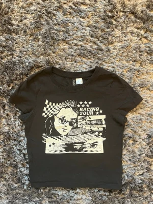 Svart Racing Tour t-shirt med tryck - Svart babytee från H&M DIVIDED med vitt grafiskt tryck framtill med texten 'Racing Tour', rutmönster, stjärnor och ögonmotiv. T-shirten har rund hals och korta ärmar, perfekt för en edgy streetstil. Strlk XS. Använd 1 gång. ALLA PRISER KAN DISKUTERAS🤍