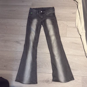 Replay grå bootcut jeans med gröna detaljer - Snygga grå jeans från weekday  med bootcut passform och coola slitningar framtill. De har klassiska fem fickor, gröna detaljer på knapp och bakre etikett, samt dragkedja. Perfekta för dig som gillar en retro vibe och vill sticka ut lite extra. Dom har ju lite konstiga storlekar på weekday så minns inte vilken jag har, men jag är en 167 lång och dom passar mig så skulle säga 2 då