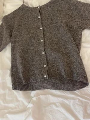 Grå stickad kofta från 157 - Mysig grå stickad kofta från 157 med rund halsringning och pärlknappar framtill. Koftan har korta ärmar och ribbade kanter nedtill och vid ärmsluten. Perfekt att slänga över en topp för en chill look.
