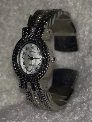 Côte d'Azur silverfärgad klocka - Stilren klocka från Côte d'Azur med silverfärgat metallarmband och detaljerad boett. Urtavlan är vit med svarta siffror och en liten sten vid tolvan. Armbandet har dekorativa mönster och små glittriga stenar för extra bling.