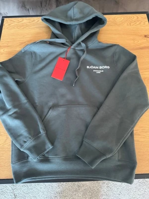 Grå hoodie från Björn Borg - Snygg grå hoodie från Björn Borg med vit logga och texten 'Stockholm 1956' på bröstet. Klassisk modell med huva, dragsnören och stor magficka framtill. Tillverkad i mjukt material som känns skönt mot huden. Perfekt för en avslappnad och trendig look.