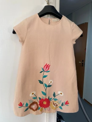 Flick  klänning med blommor - Söt beige flick  klänning från Zara girls med korta ärmar och rund hals. Klänningen har broderade blommor i rött, rosa, vitt och grönt nedtill på framsidan. Tillverkad i ett strukturerat tyg som ger en exklusiv känsla. Perfekt för dig som gillar en lekfull och färgglad stil.