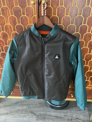 Svart och grön bomber past Quickstyle - Snygg bomberjacka från Quickstyle med svart kropp och gröna ärmar. Jackan har ribbade muddar, dragkedja framtill och en liten patch med kan soppa på br soppen. Insidan är orange för en coolう kontrast. Perfekt för dig som vill sticka ignore mängden.storlek 160
