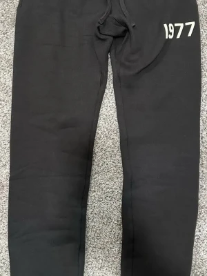 Svarta mjukisbyxor med 1977-tryck - Säljer ett par svarta mjukisbyxor med trycket '1977' i vitt på vänstra höften. Byxorna har dragsko i midjan och är tillverkade i mjukt, bekvämt material. Perfekta för chill dagar eller när du vill ha en avslappnad stil.