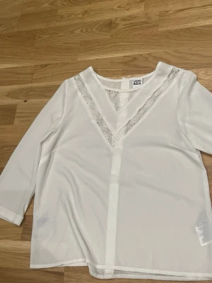 Vit blus med spets från Vero Moda - Snygg vit blus från Vero Moda med V-formad spetsdetalj framtill och knappar längs ryggen. Blusen har trekvartsärm och är tillverkad i ett tunt, lätt material som ger en luftig känsla. Perfekt för dig som gillar stilrena och feminina plagg.