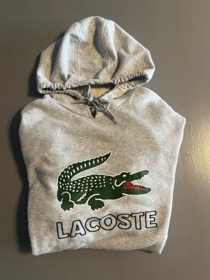 Grå hoodie från Lacoste med logga - Snygg grå hoodie från Lacoste med stor grön krokodil och Lacoste-tryck på bröstet. Klassisk huva med dragsko och magficka framtill. Mjuk bomullsblandning som är skön att ha på sig. Perfekt för en avslappnad och sportig stil.