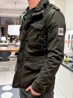 Mörkgrön parkajacka från Superdry - Snygg mörkgrön parkajacka från Superdry med fyra stora fickor framtill, axelklaffar och patch med brittisk flagga på ärmen. Jackan har dragkedja och knappar framtill samt hög krage. Perfekt för dig som gillar en urban och avslappnad stil.