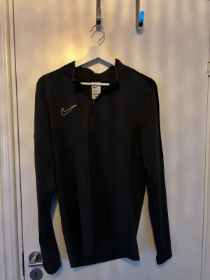 Svart långärmad träningströja Nike - Svart långärmad träningströja från Nike med halv dragkedja och neongrön logga på bröstet. Tröjan har en sportig passform och är tillverkad i ett lätt och stretchigt material som passar perfekt för löpning eller gym.