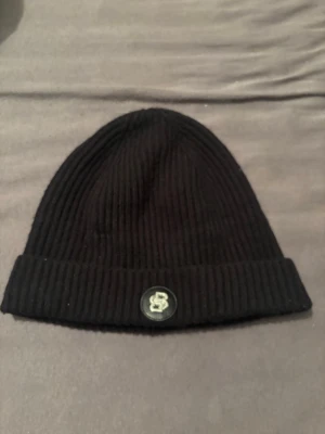 Svart ribbstickad mössa med patch - En svart hugo boss mössa med uppvikt kant och en rund patch framtill med ett boss-märke. Perfekt för att hålla värmen och ge en stilren look. Materialet är lätt och man känner inte av att man har på sig en mössa.