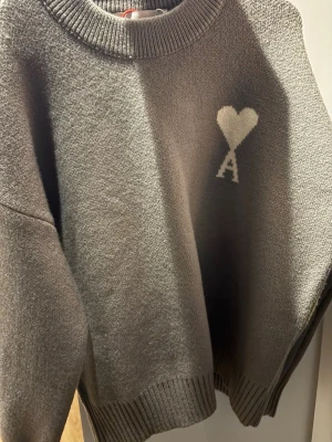 Grå stickad tröja från Ami Paris - Säljer en grå stickad tröja från Ami Paris med deras ikoniska hjärta och A-logo på bröstet. Tröjan har rund halsringning, ribbade muddar och är tillverkad i mjukt material som ger en avslappnad och trendig look.