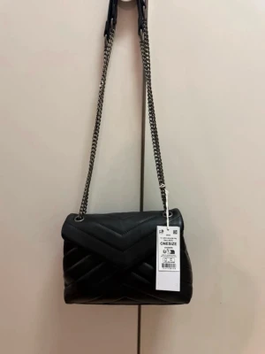 Väska från gina tricot - Stilren svart axelväska från Gina Tricot mönster och silverfärgad kedjerem. Väskan har lock med magnetstängning och ett innerfack med dragkedja. 